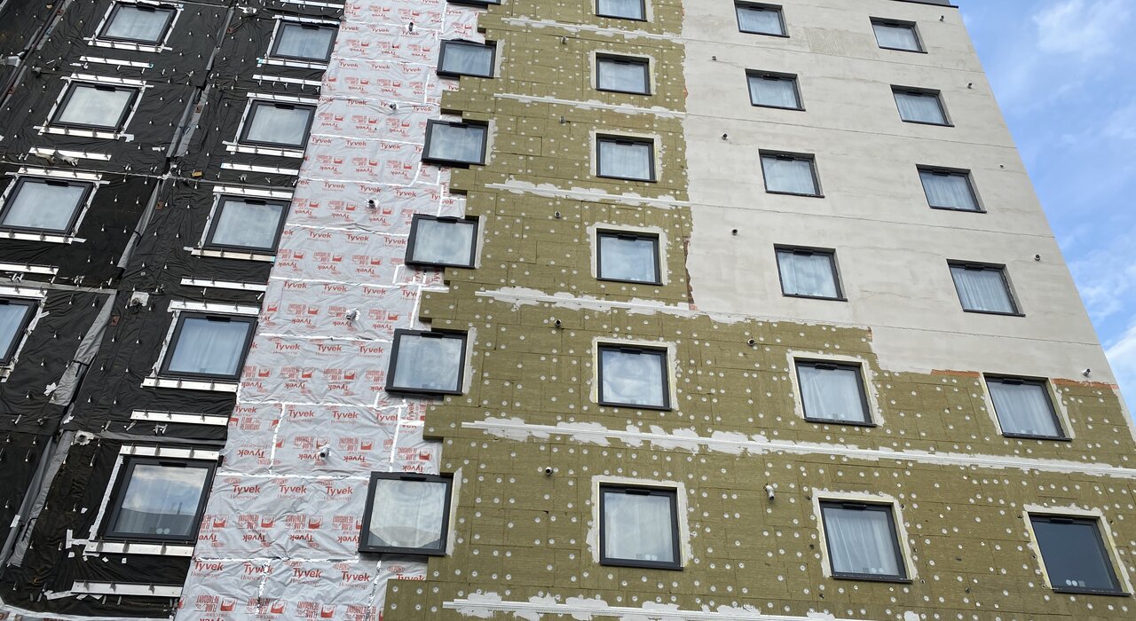 This image shows the installation process of an external wall insulation (EWI) system on a multi-storey building. The different layers—including insulation boards, weatherproof membranes, and base coats—are visible, demonstrating the step-by-step construction that improves thermal efficiency and building protection.