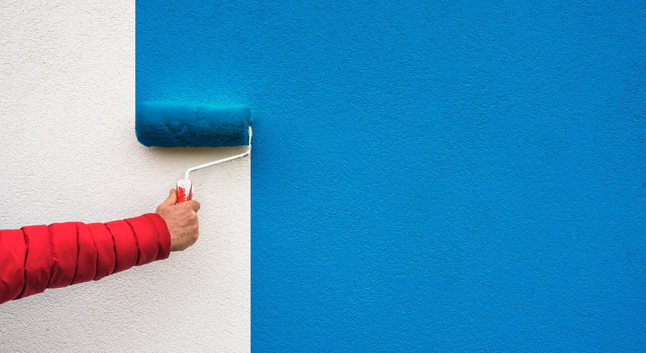 A hand uses a paint roller to apply vibrant blue paint to a white textured wall, creating a sharp contrast. This type of exterior paint finish can be used on insulated walls to add a protective, decorative coating, often as the final step in an external wall insulation system.
