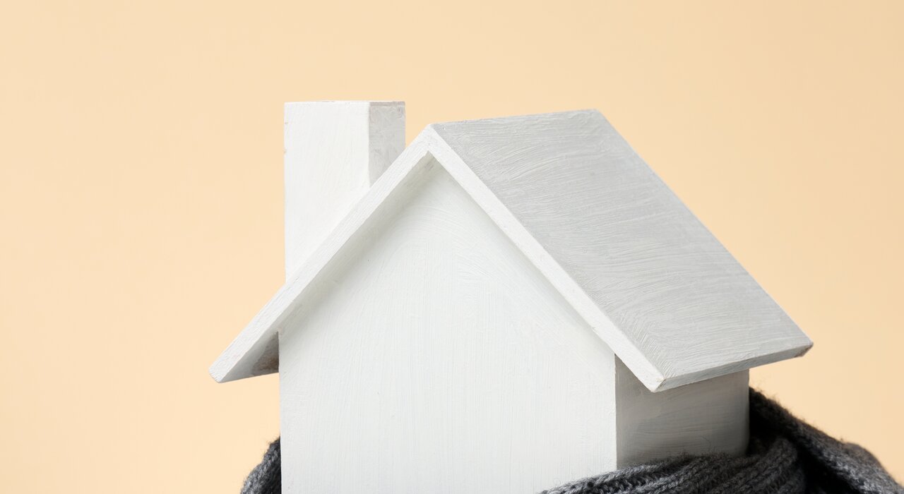 Small white wooden house model wrapped in a grey knitted scarf, symbolising home insulation and warmth, set against a soft beige background.