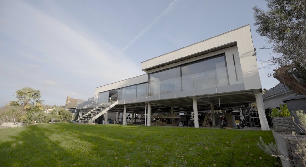  Modern elevated home with large glass panels and a smooth rendered exterior, likely finished with an external wall insulation system for optimal energy efficiency. EWI Store supplies render and insulation solutions ideal for contemporary architecture like this.