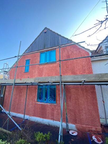 House façade under renovation, with fresh red render applied over an external wall insulation system. Windows are masked for protection, and scaffolding is in place for safe access. This render system, supplied by EWI Store, enhances energy efficiency and provides a modern, durable finish.