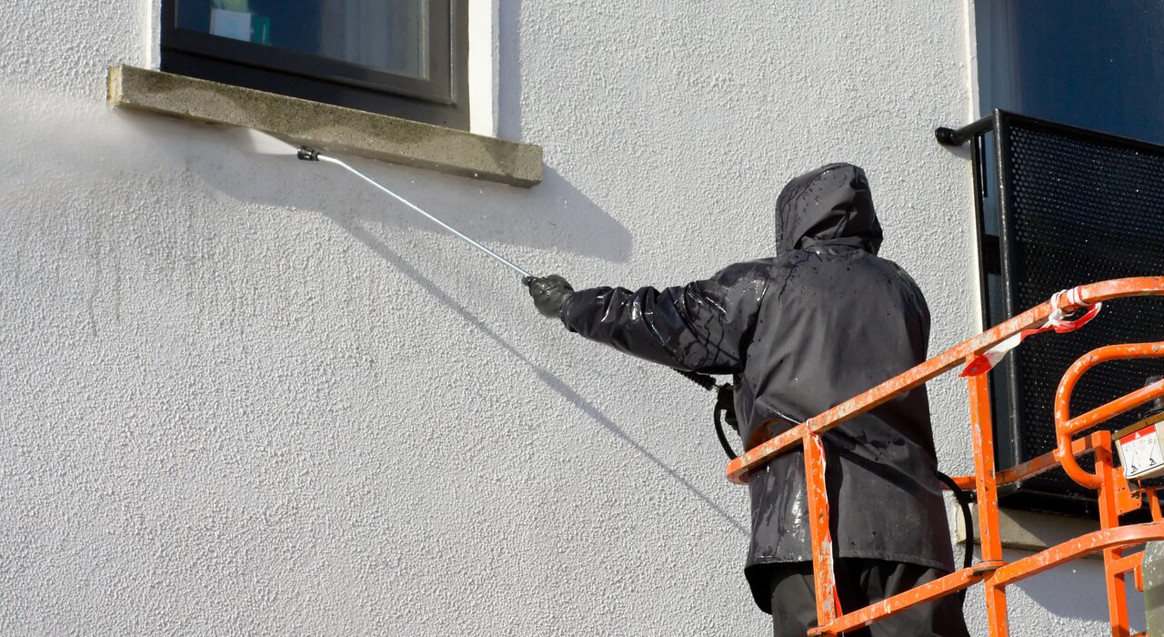 Contractor using a high-pressure washer to clean a rendered exterior wall on a building finished with an external wall insulation system. Regular maintenance like this helps preserve the appearance and performance of silicone or acrylic render finishes—available from EWI Store, specialists in render and insulation solutions.