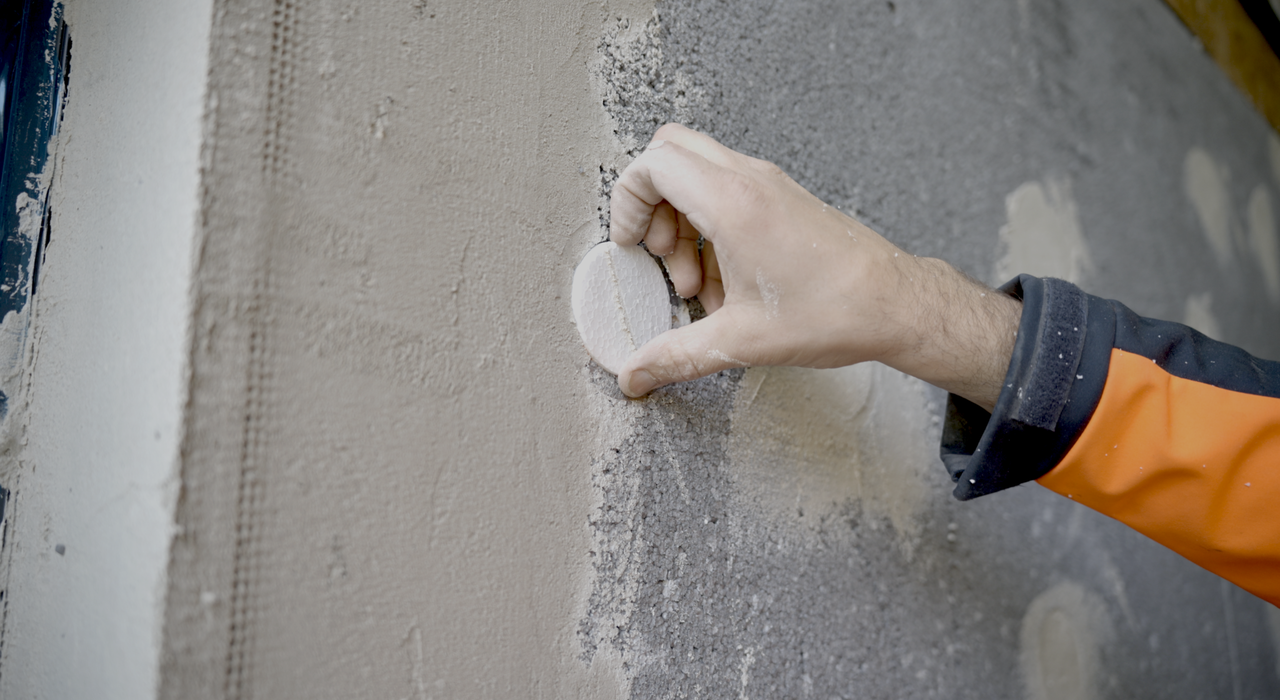 Close-up of a contractor inserting an insulation fixing cover cap into a pre-installed fixing head on a basecoated EWI system. These caps help prevent thermal bridging and ensure a smooth, uniform render finish. Available from EWI Store, they are essential components for high-performance external wall insulation installations.