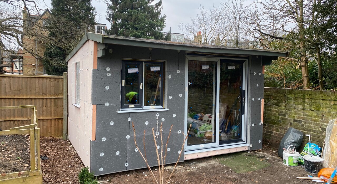 Small outbuilding under construction with grey EPS insulation boards mechanically fixed to the exterior as part of an external wall insulation system. The boards are prepared for basecoat and render application. Materials like these are available from EWI Store, providing energy-efficient solutions for garden rooms, extensions, and new builds.