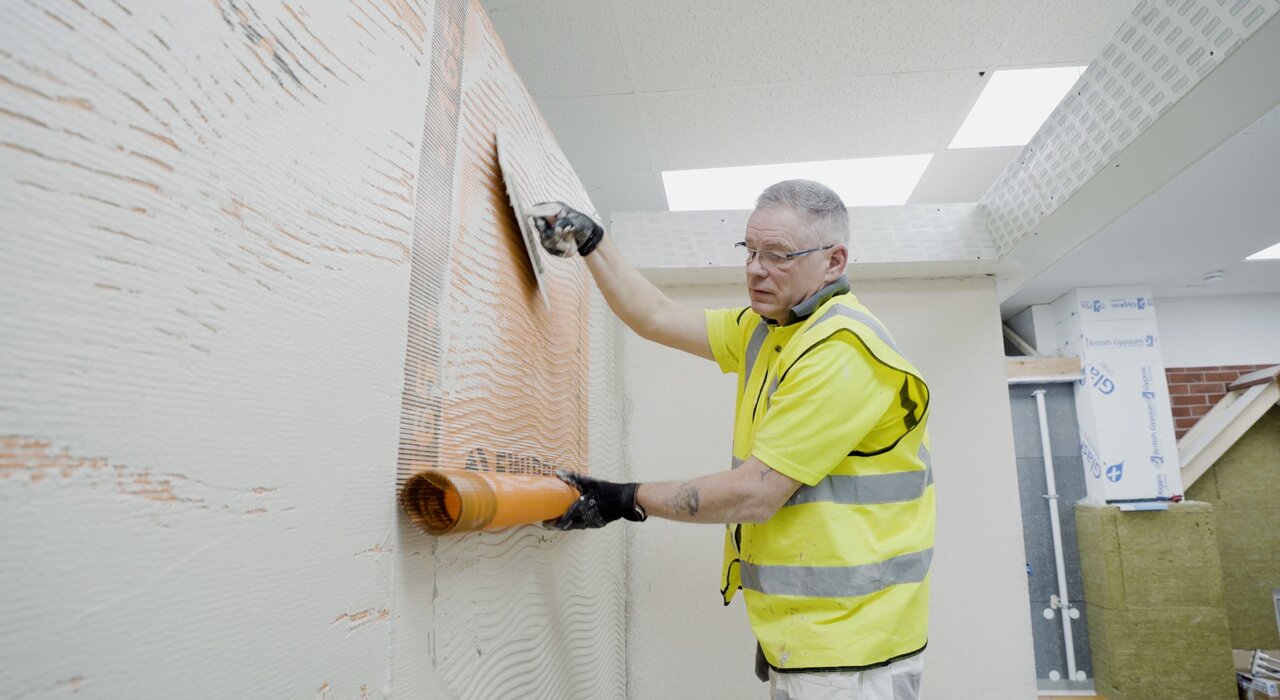 Professional installer embedding orange fibreglass mesh into a freshly applied basecoat layer on an external wall insulation system. This step reinforces the surface, preventing cracks and improving durability. Materials like mesh, basecoat, and tools used here are supplied by EWI Store for high-performance EWI applications.