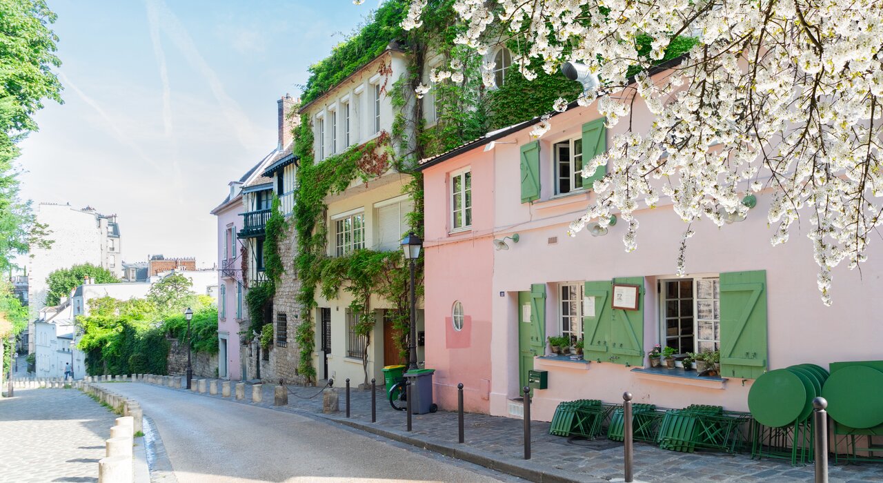 A quaint street in Montmartre, Paris, with pastel buildings, green shutters, ivy-covered walls, and blooming cherry blossoms under a clear blue sky.
