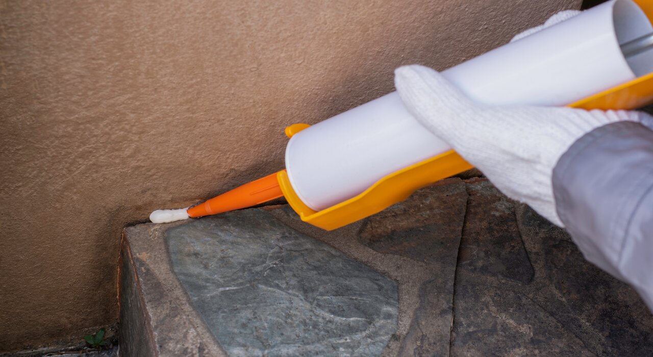 A close-up of a person applying sealant with a caulking gun along the edge of an exterior wall, preparing the surface for external wall insulation.