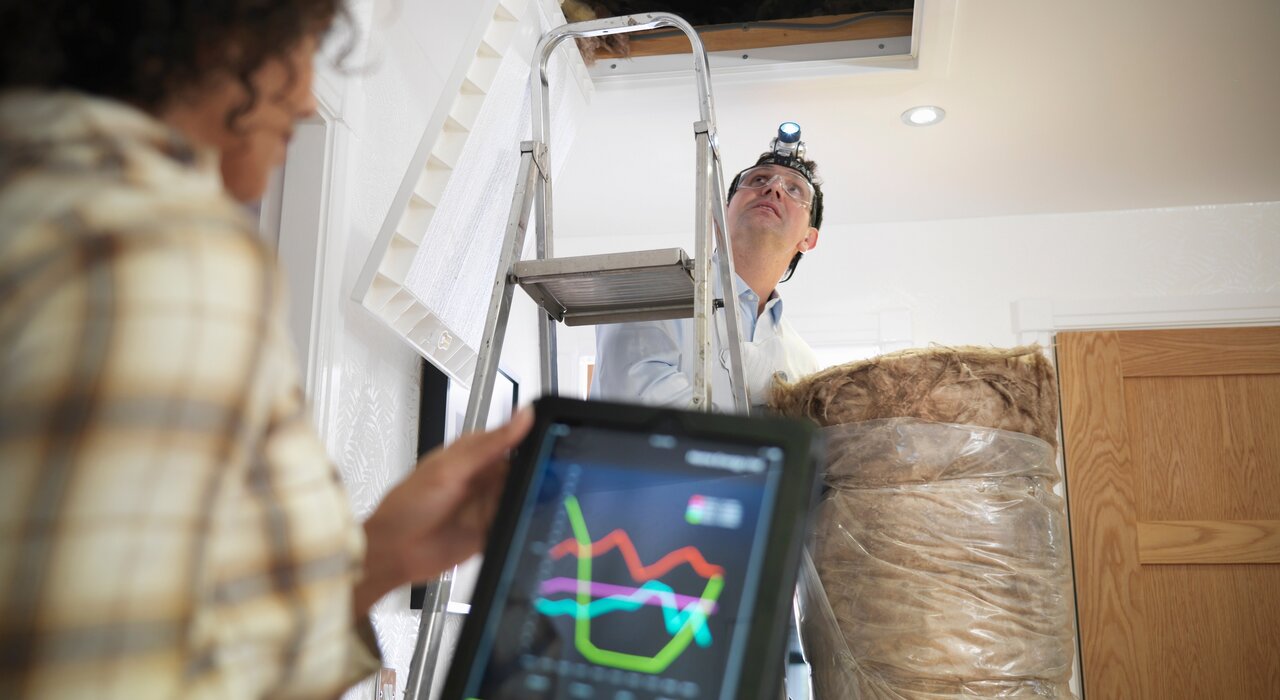 Person inspecting attic insulation with a headlamp and ladder, while another person monitors data on a tablet in the foreground.