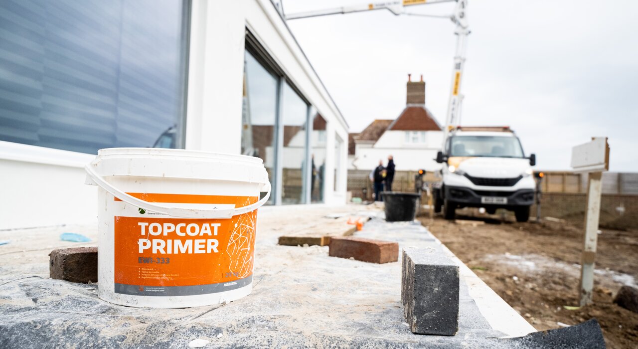 A bucket of Topcoat Primer placed on a construction site surface in front of a modern building; used as a base layer to ensure strong adhesion and durability for exterior wall coatings.