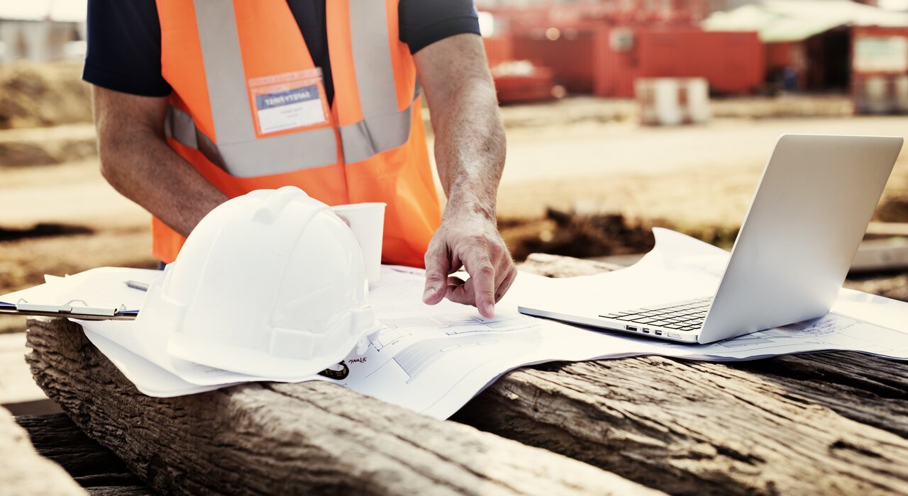 Construction site worker wearing a high-visibility orange vest pointing at blueprints spread over timber, with a white safety helmet and open laptop beside him. The background shows a blurred construction area with equipment and materials.