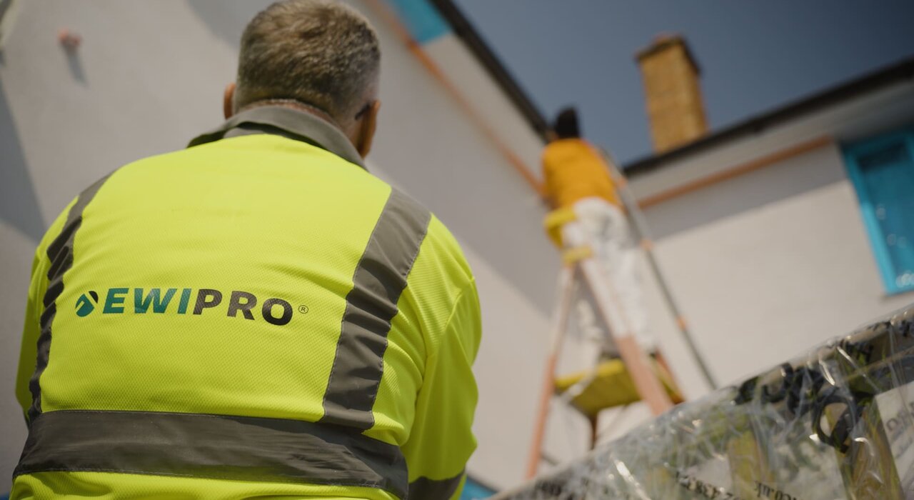 A construction worker wearing a high-visibility jacket with the "EWI PRO" logo watches another worker standing on a ladder, applying render to the exterior of a white building under renovation.