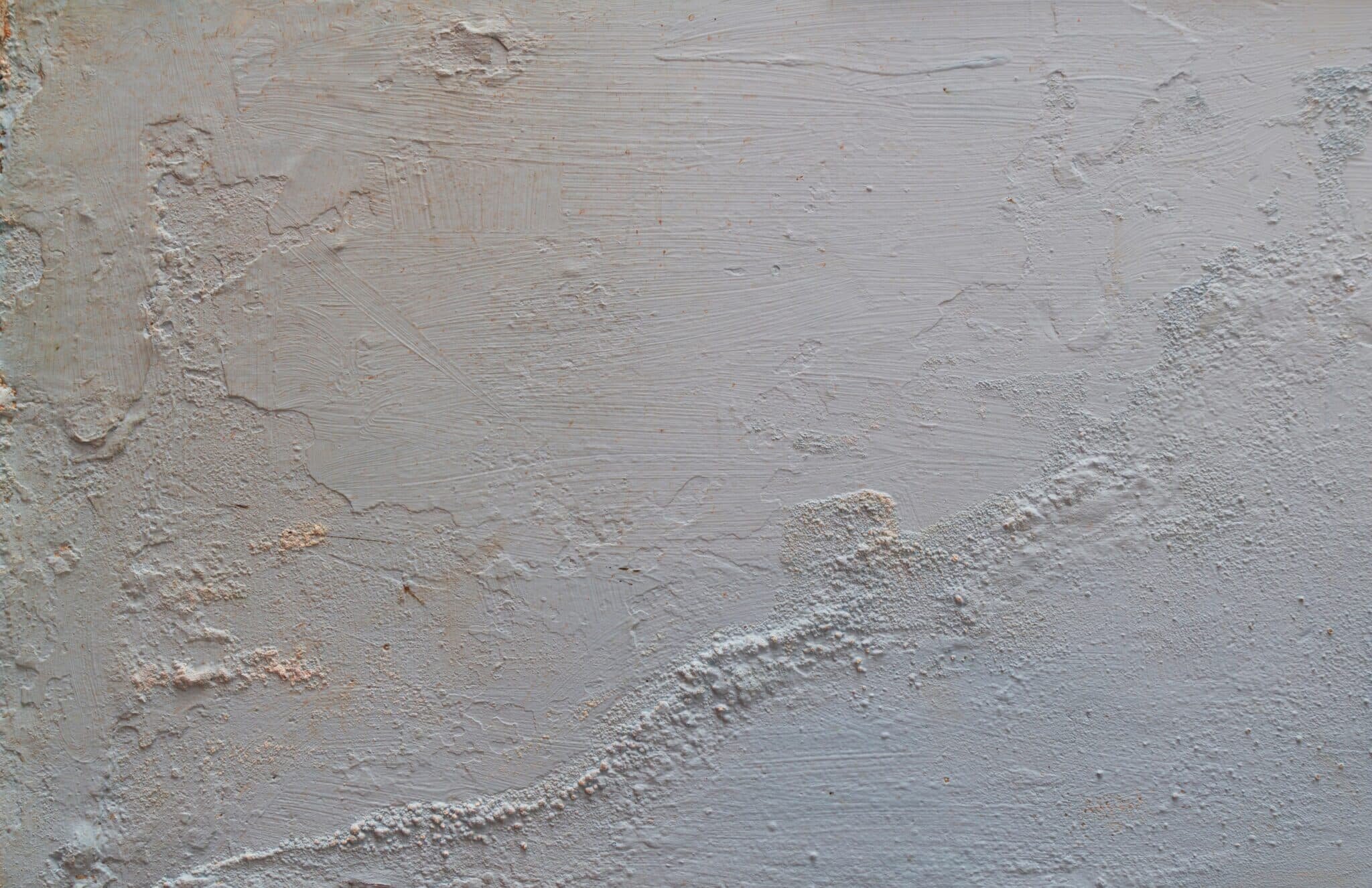 Close-up of a wall with a rough and uneven plaster surface. The wall shows signs of wear, including cracks, bumps, and patchy texture, possibly indicating moisture damage or an aging finish. The color is a muted, off-white or pale grey.