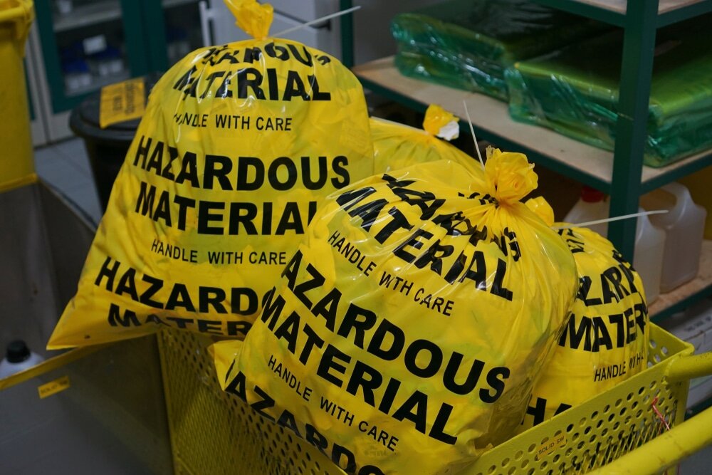 Bright yellow bags marked with bold black text stating hazardous material handle with care are filled and tied securely. The bags are placed inside a yellow plastic container in what looks like a storage or waste disposal area, with shelving units and supplies visible in the background, indicating the safe handling of potentially dangerous waste.