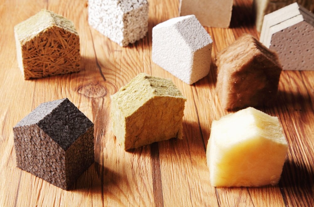 This image shows various types of insulation materials shaped like small houses, each representing different thermal insulation options used in construction, such as mineral wool, foam board, wood fibre, and natural fibre panels.