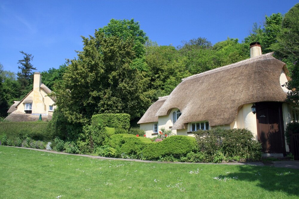 This image shows traditional thatched cottages surrounded by lush greenery and a well-maintained garden, likely in a rural or countryside setting. The thick thatched roofs help with natural insulation, keeping the homes cooler in summer and warmer in winter.