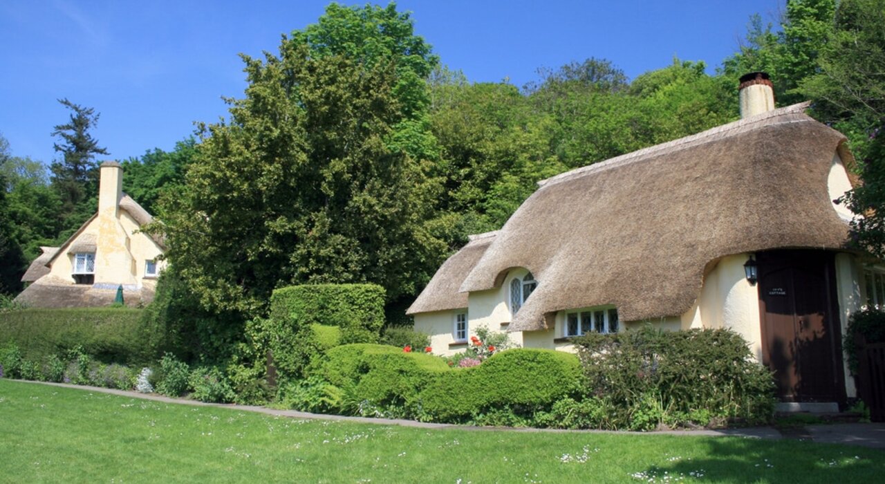 This image shows traditional thatched cottages surrounded by lush greenery and a well-maintained garden, likely in a rural or countryside setting. The thick thatched roofs help with natural insulation, keeping the homes cooler in summer and warmer in winter.