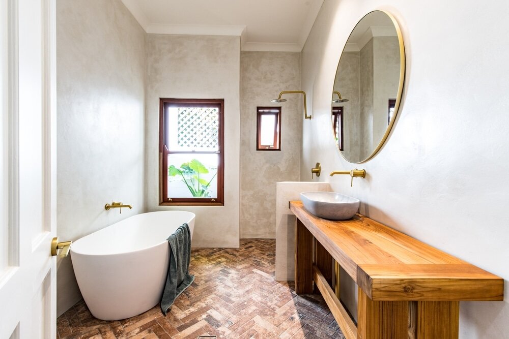 This image shows a modern, minimalist bathroom with natural and warm finishes. It features a freestanding white bathtub, brass fixtures, a wood vanity with a stone basin sink, and a round mirror. The herringbone-style flooring adds texture and complements the neutral-toned walls, while the small windows bring in natural light and a view of greenery outside.