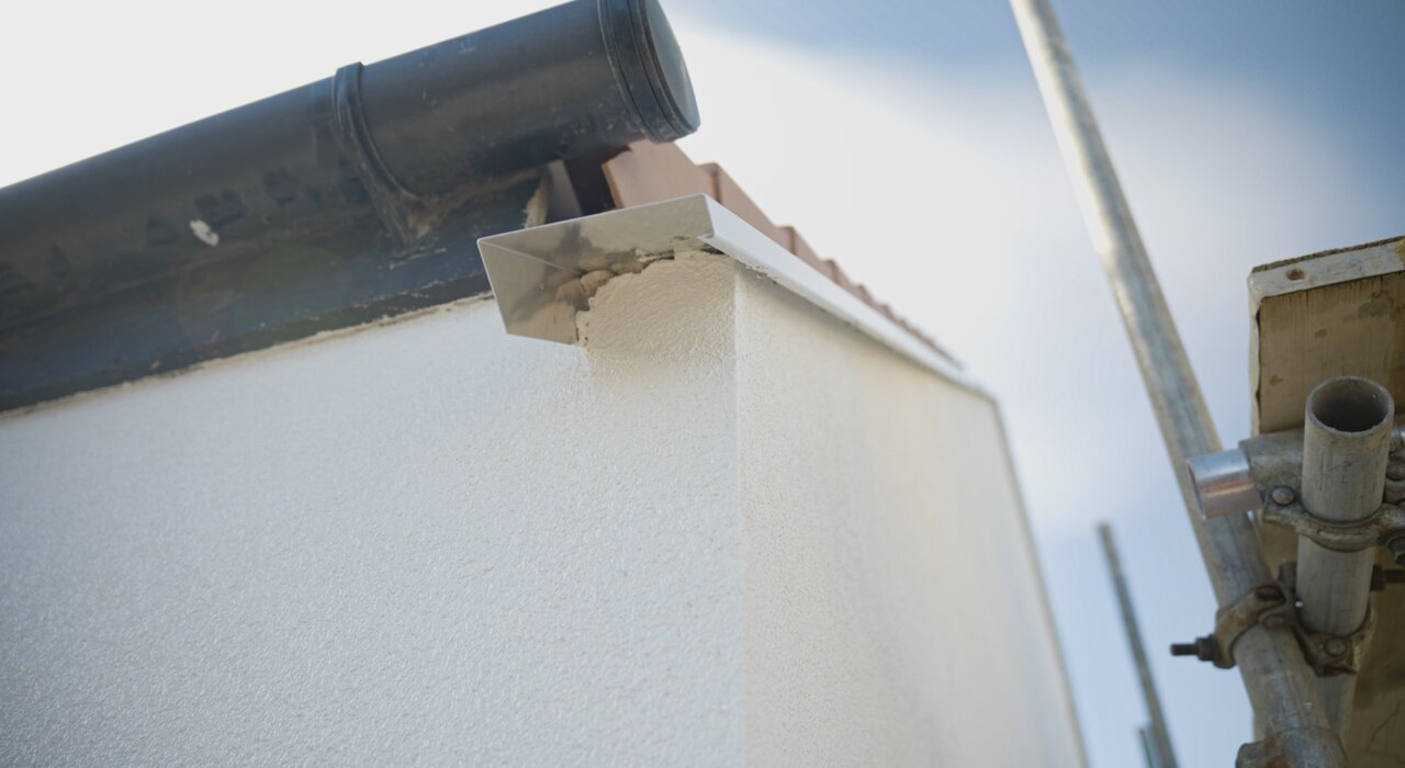 This image shows an external corner of a building fitted with external wall insulation and a render finish. A metal verge trim is installed to cap and protect the top edge where the insulation meets the roof tiles and guttering, preventing water ingress and maintaining a clean, finished look. This detail is crucial for ensuring weather resistance and prolonging the life of the insulation system.