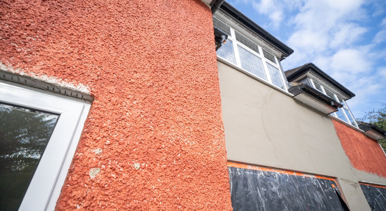  A close-up of a house wall showing two different stages of external wall insulation. The red textured surface represents the original finish, while the smoother grey section shows new EWI installation in progress