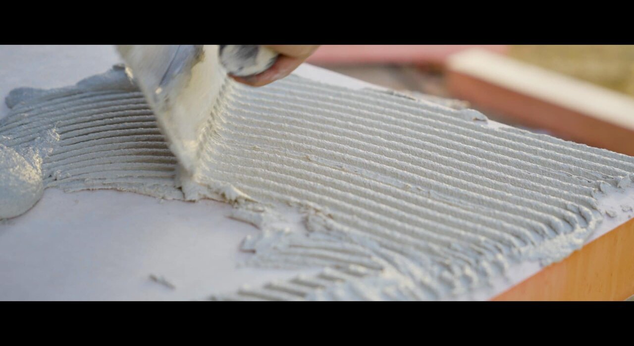 A hand is seen applying adhesive in lines across an insulation board. This is a typical step in the external wall insulation process to securely fix boards to the wall.