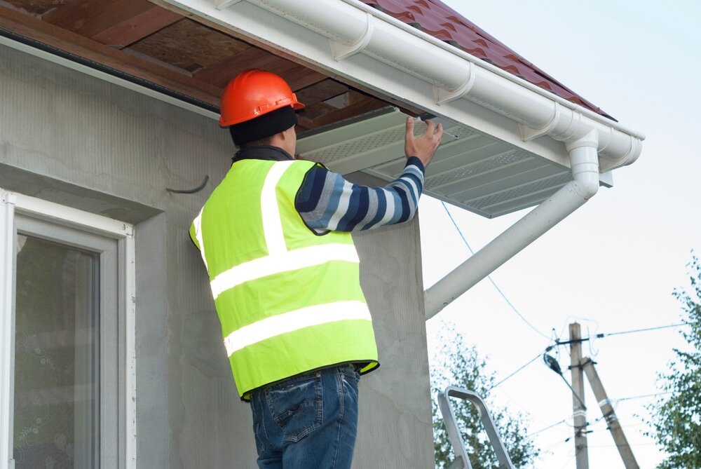 A construction worker in high-visibility clothing and a helmet inspecting or installing uPVC soffit boards under a roof overhang. Proper soffit and fascia installation plays a key role in ventilation, moisture protection, and neat finishing on EWI systems.