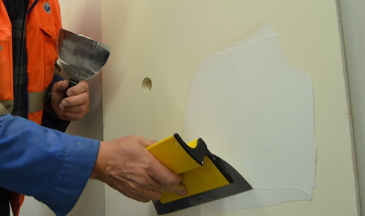 A worker is applying a thin layer of skim coat or filler to a plasterboard wall using a wide scraper, smoothing out imperfections. This process is essential for patch repairs or preparing walls for decoration, especially after insulation upgrades or surface damage.