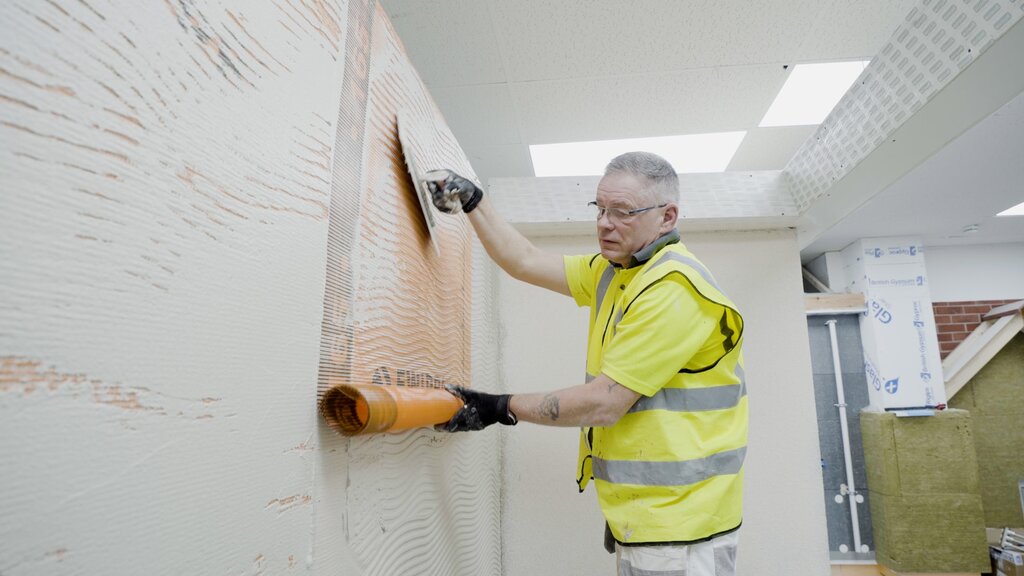 A skilled installer embeds orange fibreglass mesh into a fresh basecoat layer on an insulated wall. This reinforcement step is crucial in EWI systems to prevent cracking and improve durability before applying the final render. The materials are supplied by EWI Store.