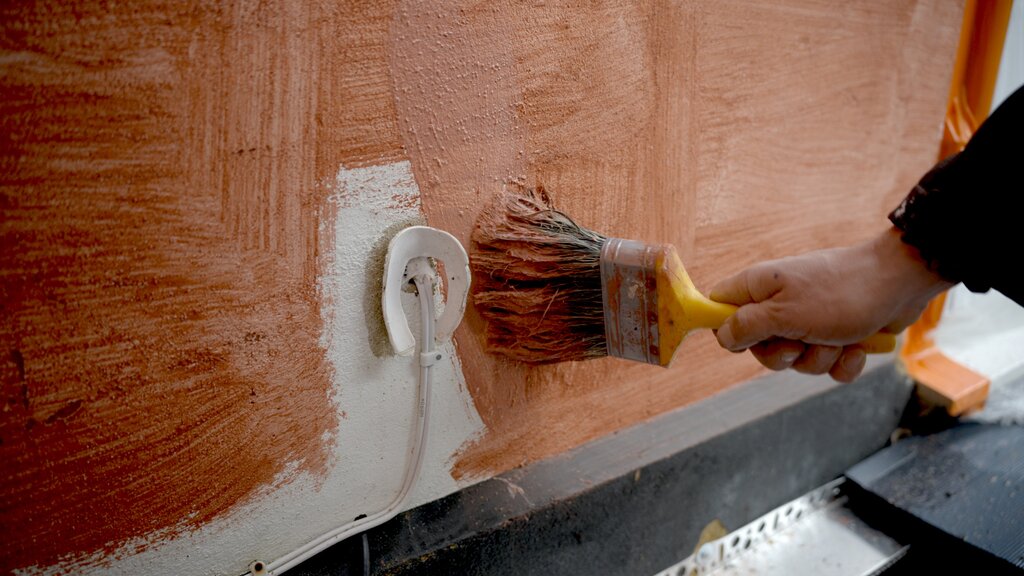 Close-up of a worker brushing on red-tinted primer over a cured basecoat layer, carefully working around a wall outlet. Priming ensures proper adhesion of the finishing render layer. EWI Store offers a range of primers suited to silicone, acrylic, or mineral renders.