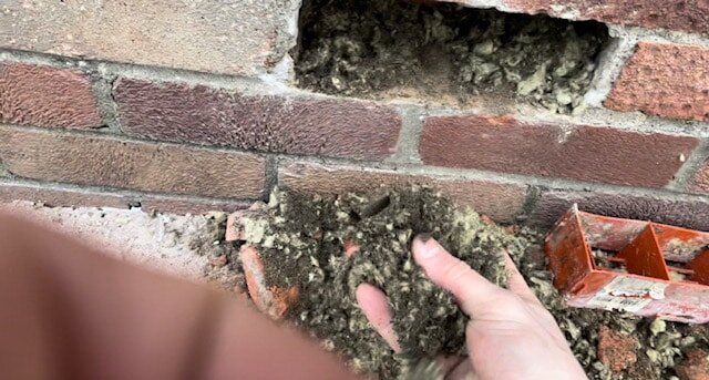Close-up of a hand removing degraded cavity wall insulation from a brick wall. This image highlights the importance of proper insulation removal prior to retrofit works. EWI Store provides expert advice and products for safe extraction and system preparation.