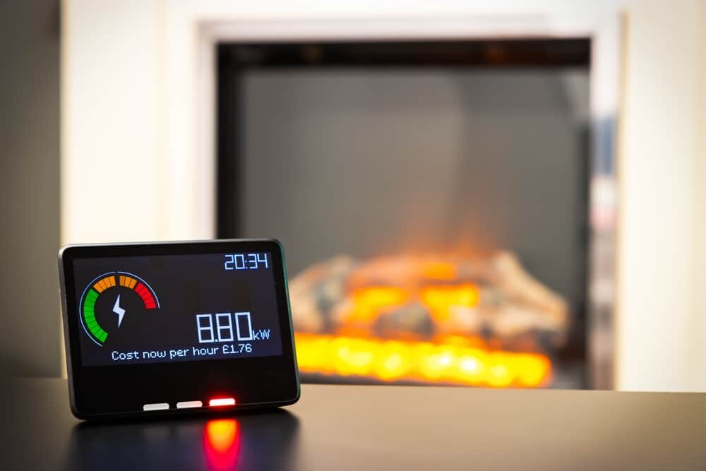 Close-up of a smart energy monitor displaying real-time electricity usage, positioned in front of a lit electric fireplace. The display shows a cost of £1.76 per hour at 8.80kW, highlighting energy consumption awareness. At EWI Store, we advocate for better insulation to lower heating demands and reduce energy bills—upgrade your thermal envelope and stay in control.