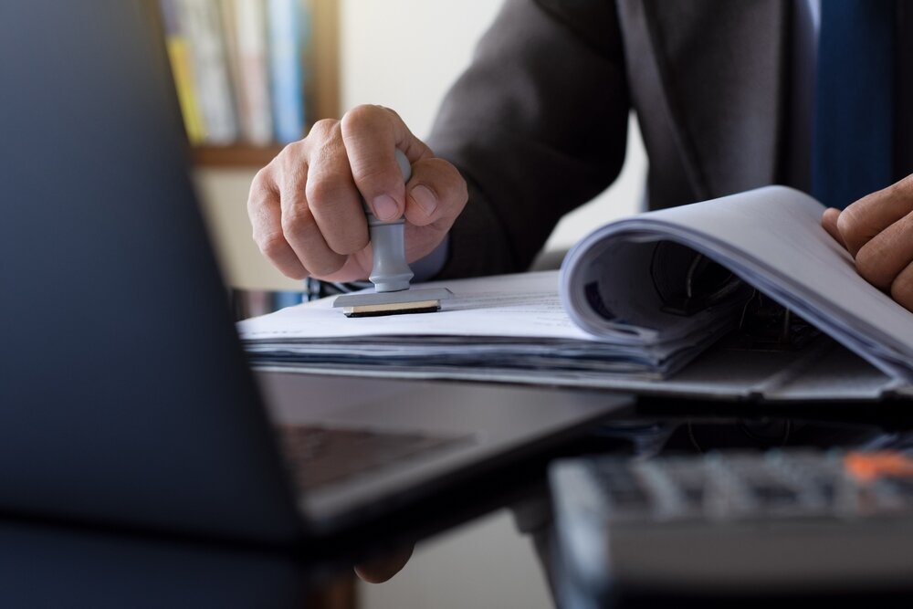 A businessperson stamping documents at a desk, likely representing planning approval or grant documentation. EWI Store offers guidance on meeting Building Regulations and accessing government funding for energy efficiency upgrades, including insulation and render systems.