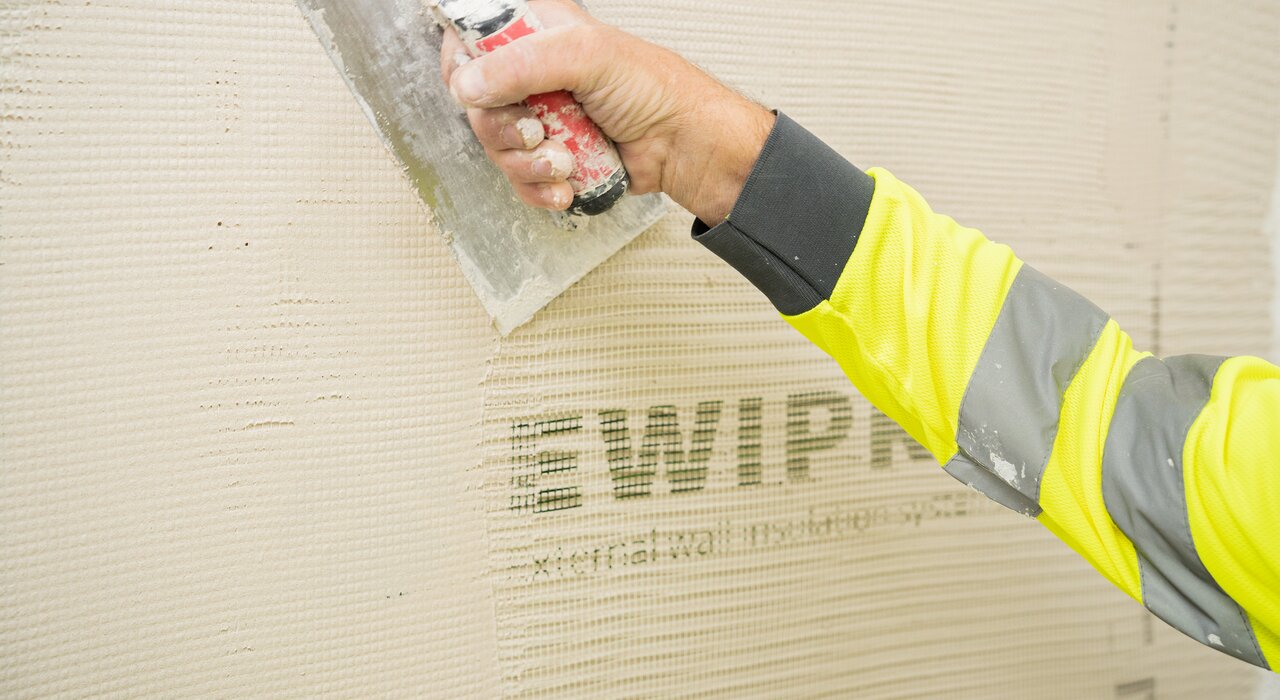 Close-up of a worker applying mesh-embedded basecoat using a stainless steel trowel on external wall insulation. The visible EWI Pro branding on the mesh indicates use of a certified and reliable system, available from EWI Store for effective thermal performance and surface reinforcement.