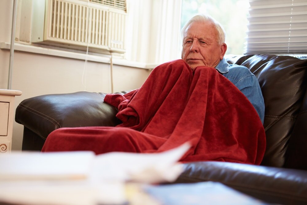 Elderly man wrapped in a blanket on a sofa, visibly cold inside his home. A stark reminder of the importance of proper insulation and heating efficiency—problems that EWI Store solutions like EWI systems and breathable renders are designed to solve.