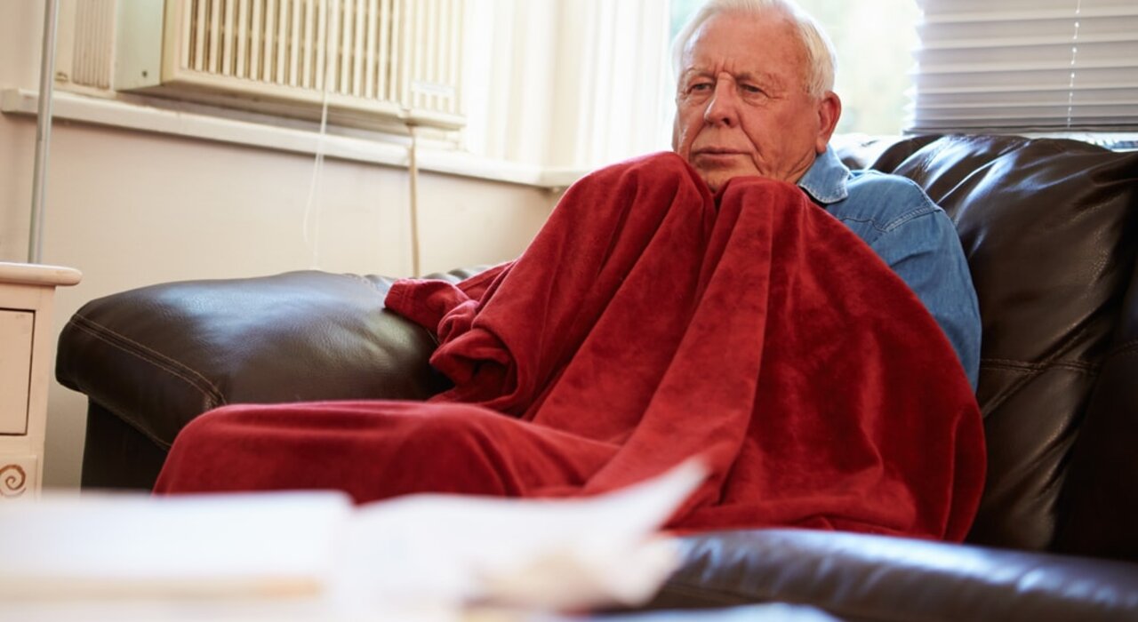 Elderly man wrapped in a blanket on a sofa, visibly cold inside his home. A stark reminder of the importance of proper insulation and heating efficiency—problems that EWI Store solutions like EWI systems and breathable renders are designed to solve.