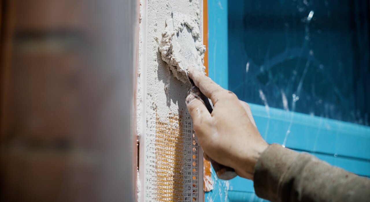 Worker applying basecoat over fibreglass mesh at a wall corner. The mesh is embedded in a thick, smooth layer for reinforcement and crack resistance. EWI Store supplies complete render systems including mesh, basecoats, and corner profiles for durable façade finishes.