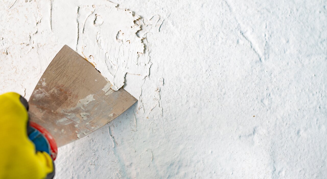 Close-up of a worker removing damaged or flaking paint from a rendered surface using a metal scraper. Proper surface preparation is essential for achieving long-lasting results when applying EWI Pro renders and paints. EWI Store offers a full range of primers, tools, and finishing products to ensure professional-grade external wall insulation installations.