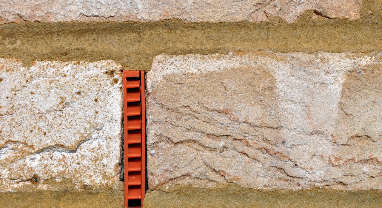 Close-up of a red plastic wall weep vent installed in the mortar joint of a stone wall. These weep vents are essential for cavity wall drainage and ventilation, preventing moisture build-up and damp issues. Available at EWI Store, our ventilation accessories support a healthy building envelope and effective moisture management.